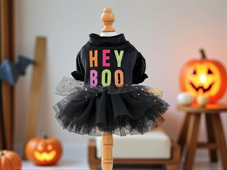 May include: A black pet dress featuring a mock neck and short sleeves. The bodice displays the text HEY BOO in colorful, glittery letters. The skirt consists of layered black tulle fabric adorned with small, shimmering gold star-shaped sequins.