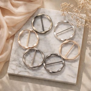Op de afbeelding: Zes metalen sjaalringen in goud, zilver en brons. Elke ring heeft een gedraaid cirkelvormig ontwerp met een rechte staaf in het midden. De ringen zijn gerangschikt op een marmeren oppervlak, met een zachte, neutraal gekleurde stof op de achtergrond.