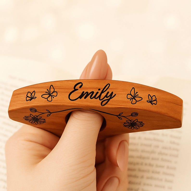 May include: A wooden thumb page holder with the name "Emily" in black script. The holder is decorated with butterfly and floral designs. The thumb hole is visible, and the holder is held by a hand.