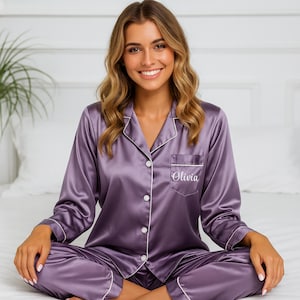 Puede incluir: Un conjunto de pijama de satén lavanda con ribete blanco y el nombre "Olivia" bordado en el bolsillo. El conjunto incluye una camisa con botones y pantalones a juego. La modelo está sentada en una cama.
