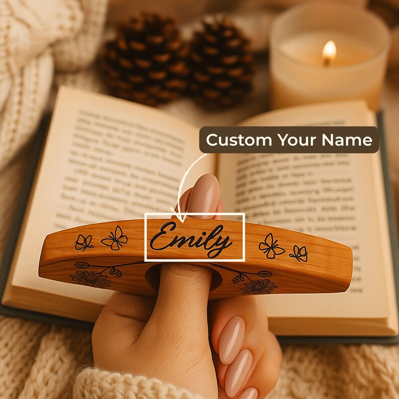 May include: A wooden book page holder with the name "Emily" engraved on it. The holder features floral and butterfly designs. The text "Custom Your Name" is also visible. The holder is being used on an open book.