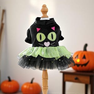 May include: A black pet dress featuring a cat face design with large green eyes, pink triangular ears, and a pink heart-shaped nose. The skirt consists of layered lime green and black tulle fabric adorned with small gold star-shaped sequins.