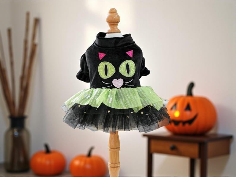 May include: A black pet dress featuring a cat face design with large green eyes, pink triangular ears, and a pink heart-shaped nose. The skirt consists of layered lime green and black tulle fabric adorned with small gold star-shaped sequins.