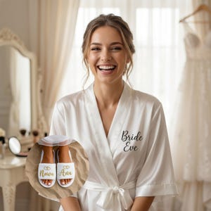 Może przedstawiać: Uśmiechnięta osoba w białym satynowym szlafroku z napisem "Bride Eve" czarnym pismem. Na zdjęciu widoczne są również białe kapcie ze złotym napisem "Nowa Ana". Szlafrok ma wiązanie.
