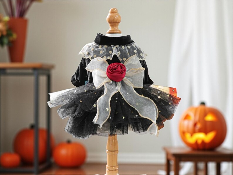 May include: A black velvet pet dress featuring a layered tulle skirt with gold star accents, a high collar, and a large sheer white bow at the waist centered with a deep red fabric rose. The garment is displayed on a wooden mannequin stand.