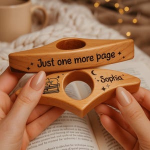 May include: Two wooden book page holders. One has the text "Just one more page" and the other has the name "Sophia". Both have a circular opening and star and moon designs. The holders are light brown and are held over an open book.