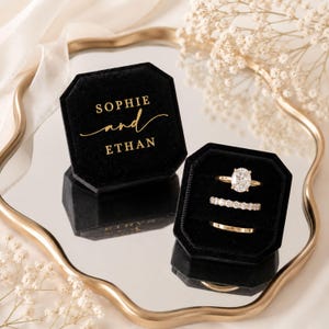 Può includere: Due scatole ottagonali per anelli in velluto nero con scritte dorate SOPHIE and ETHAN. Una scatola è chiusa, l'altra è aperta e mostra tre anelli d'oro: un solitario ovale, una fede con diamanti e una fede nuziale in oro semplice.
