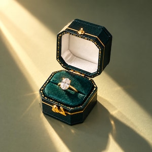 Op de afbeelding: Een solitaire ring met een ovaal geslepen diamant en een gouden band bevindt zich in een achthoekig donkergroen fluwelen juwelendoosje. Het doosje is voorzien van gouden randen, een witte binnenvoering en een gouden metalen sluiting.