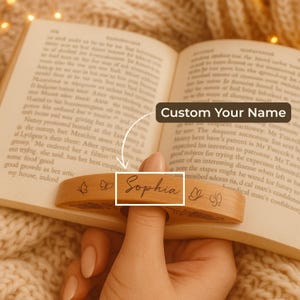 May include: A wooden book holder with the name "Sophia" engraved on it. The holder is light brown and features butterfly designs. It is designed to hold a book open while reading.