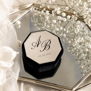 May include: A black velvet hexagonal ring box with a mirrored lid featuring the engraved initials AB and the date 06.22.2027 in a black script font. The box has a sleek, geometric shape and is designed to hold jewellery securely.