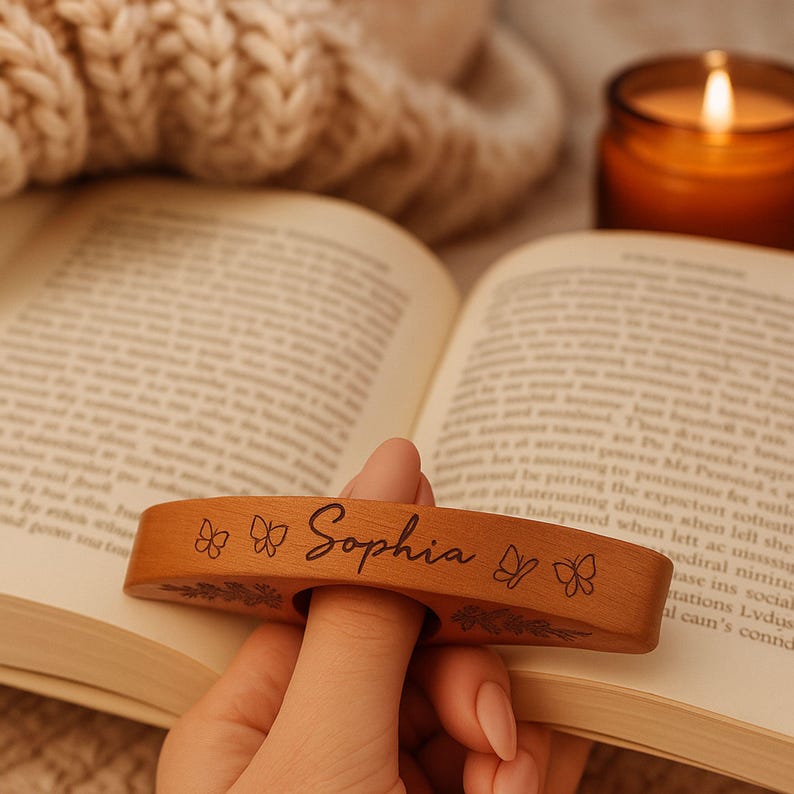 May include: A wooden thumb page holder with the name "Sophia" engraved on it, along with butterfly and floral designs. The page holder is resting on an open book, with a lit candle in the background.