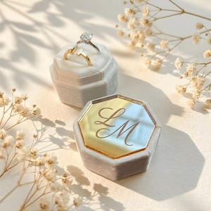 May include: Two engagement rings rest in a light gray velvet ring box. One ring has a large, clear gemstone, and the other is gold. A second ring box with a gold mirrored top displays the initials "LM".