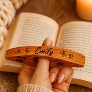 May include: Wooden book page holder with the name "Emily" in black script. The holder is decorated with floral and butterfly designs. The holder is held over an open book.