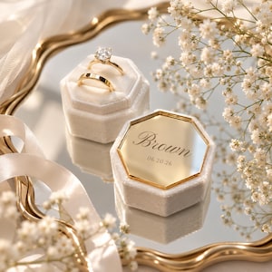 Puede incluir: Dos cajas hexagonales de terciopelo crema para anillos, una con un anillo de diamante solitario y una banda de oro, la otra con una tapa de espejo dorada grabada con el nombre Brown y la fecha 06.20.26, sobre una bandeja de espejo dorada.