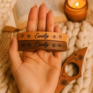 May include: Two wooden book page holders, one light and one dark, engraved with the name "Emily" and floral designs. A wooden reading accessory with floral engraving is also visible. A lit candle and a book are in the background.