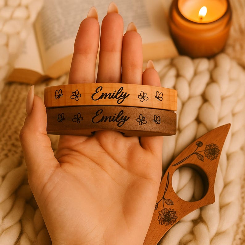 May include: Two wooden book page holders, one light and one dark, engraved with the name "Emily" and floral designs. A wooden reading accessory with floral engraving is also visible. A lit candle and a book are in the background.
