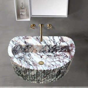 Puede incluir: Lavabo ovalado de mármol con exterior estriado. El lavabo presenta un patrón veteado blanco y burdeos, un grifo dorado y accesorios a juego. Se ve una pequeña balda con un dispensador de jabón y un espejo.