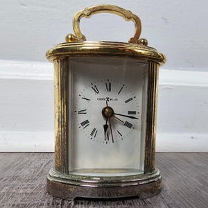 May include: A gold-toned Howard Miller quartz carriage clock with a white face, black Roman numerals, and black hands. The clock features a rectangular glass front, a metallic handle on top, and a weathered finish on the metal frame.
