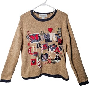 May include: A tan long-sleeve jumper with a dark blue trim at the collar, cuffs, and hem. The front features embroidered holiday-themed appliqués and the text MERRY CHRISTMAS, including a snowman, candy canes, a red heart, and gift boxes.