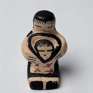May include: A small, hand-painted ceramic figurine of a parent embracing a child. The figures have black hair and are painted with black and red accents on a beige base. The base is black.