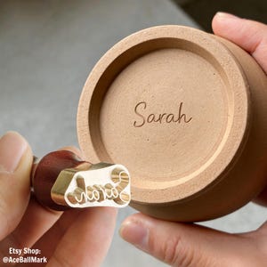 Puede incluir: Un sello personalizado con la palabra "love" en cursiva. El sello es de latón y madera, y se muestra junto a un cuenco de cerámica marrón claro con el nombre "Sarah" grabado en la base.