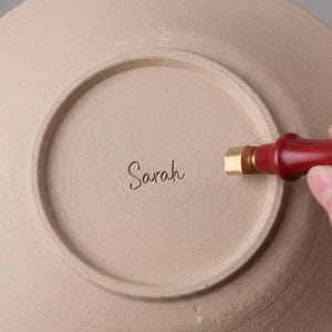 May include: A beige ceramic plate with the name "Sarah" inscribed on the bottom. A red and gold stamp is being used to mark the plate. The plate has a circular design.