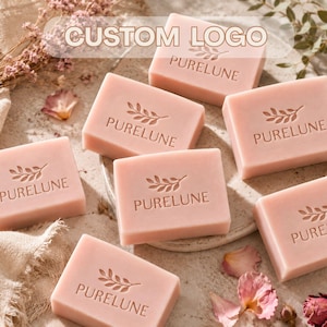 May include: Several rectangular bars of light pink soap, each embossed with a leaf icon and the text PURELUNE. The soap bars are arranged on a textured surface, accompanied by scattered dried flower petals and sprigs of greenery.