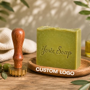 Puede incluir: Un bloque rectangular de jabón verde oliva con el texto Josie Soap grabado en el frente, apoyado sobre un pequeño plato de madera junto a un sello de latón con mango de madera. El sello presenta una base circular con borde festoneado.