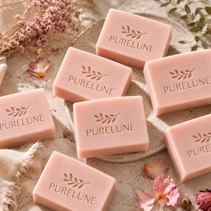May include: A collection of pink rectangular soaps, each bearing the inscription "PURELUNE" and a leaf motif, are displayed on a decorative plate. Dried flowers and petals surround the soaps, creating a spa-like atmosphere.