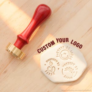 May include: A red wooden handle stamp with a brass head positioned next to a white clay disc. The clay features three distinct circular impressions with the text POTTERY BY SARAH, including a smiley face, a plant, and a sprig design. Text reads CUSTOM YOUR LOGO.