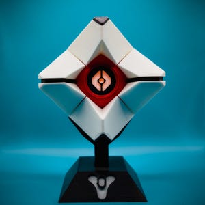 May include: A white and red geometric sculpture with a black base. The sculpture has a diamond shape with a red center and a black stand. The base has a white symbol on it. The background is a solid teal color.