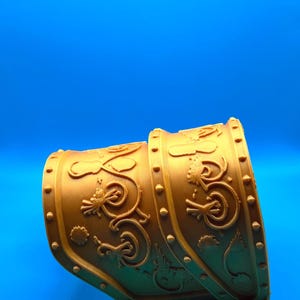 Puede incluir: Una pieza de armadura dorada y curvada con intrincados diseños en relieve de dragones y flores. La pieza tiene forma cilíndrica con remaches decorativos a lo largo de los bordes, sobre un fondo azul. Este artículo es probablemente parte de un disfraz o recreación histórica.