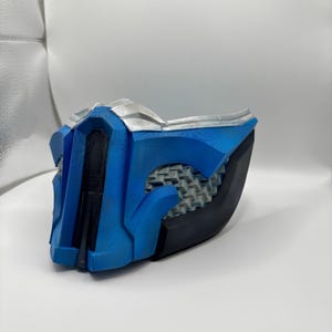 May include: A blue and black face mask featuring a central vertical ridge, a silver-colored top trim, and a textured gray mesh-like pattern on the sides. The mask has a rigid, angular construction with a smooth finish on the blue sections.