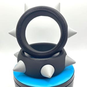 May include: Two black and grey spiked bracelets, one resting atop the other. The bracelets have a series of grey, cone-shaped spikes around the outer edge. The bracelets are on a blue and black surface.