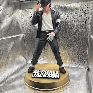 May include: A Michael Jackson figurine wearing a black fedora, a sequined black jacket, a white V-neck shirt, black trousers, and a single white glove. The figure stands on a circular gold base with the text MICHAEL JACKSON in white letters.