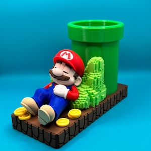 Op de afbeelding: Een 3D-geprinte figuur van een slapende Mario, met een rode hoed en blauwe overall. Hij ligt op een bruin platform met gouden munten, naast een groene pijp en gepixelde groene struiken. De achtergrond is turquoise.