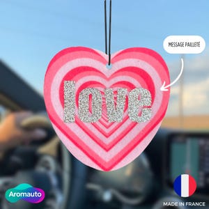May include: Heart-shaped car air freshener with a pink and red striped design. The word "LOVE" is spelled out in silver glitter. The air freshener hangs from two black strings.