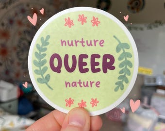 May include: A round sticker with the words "nurture QUEER nature" in purple text on a light green background. The sticker is decorated with floral and leaf accents in green and pink. Small pink hearts are scattered around the sticker.