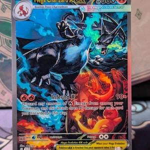 Könnte beinhalten: Eine holografische Mega Charizard X EX Pokémon-Karte. Die Karte zeigt eine dynamische Illustration von Charizard in Blau- und Schwarztönen mit feurigen orangefarbenen Akzenten. Der Kartentext enthält "Inferno X" und "90x".