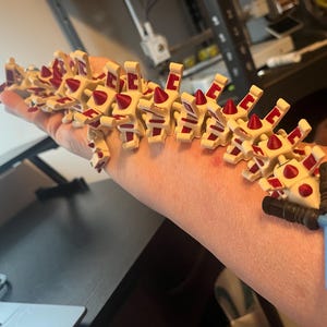 May include: A cream-colored, articulated toy with red accents and a series of connected pieces. The toy is held on a person's arm, with a black and blue toy lightsaber attached.