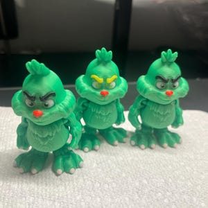 May include: Three green figurines of a grumpy character with a red nose, furrowed brow, and a tuft of hair. The figurines have a textured appearance and are standing on a white surface. The figurines are approximately 4 inches tall.