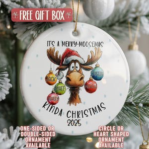 May include: A white Christmas ornament with a cartoon moose wearing a Santa hat. The ornament has the text "It's a Merry-Moosemas Kinda Christmas 2025". The moose has ornaments with names on them. Snowflake accents.
