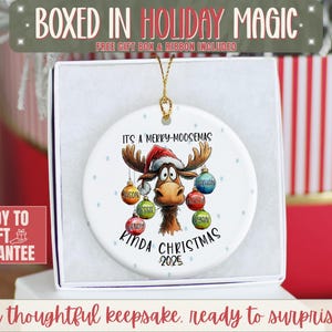 May include: A white ceramic Christmas ornament featuring a cartoon moose wearing a Santa hat. The ornament reads "It's a Merry-Moosemas Kinda Christmas 2025" with personalized names on the hanging ornaments. The ornament is in a gift box.