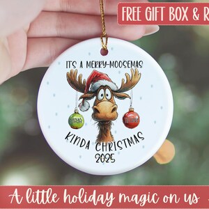 May include: A white ceramic Christmas ornament featuring a cartoon moose wearing a Santa hat. The ornament has the text "IT'S A MERRY-MOOSEMAS KINDA CHRISTMAS 2025" and two hanging ornaments with the names "MAYA" and "REBECCA".