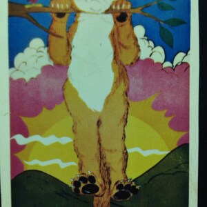 Op de afbeelding: Een vintage print met een cartoonkat die aan een tak hangt, met een kleine rode vogel. De kat heeft oranje en witte vacht, en de achtergrond bevat een zonsondergang en de tekst "Hang In There, Baby."