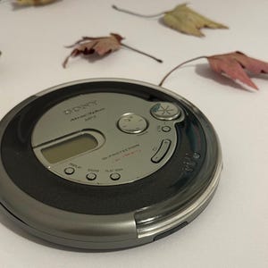 May include: A circular silver and black Sony Walkman CD player featuring an Atrac3plus MP3 logo, a small digital display, G-Protection technology, and various control buttons including display, sound, and play mode, with a volume dial.