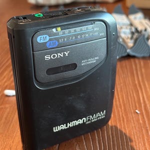 May include: A black Sony Walkman WM-FX101 portable cassette player featuring an AM/FM radio tuner with a frequency display, a volume control wheel, an anti-rolling mechanism, and a headphone jack. The device has white text branding on the front.