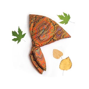 May include: A triangular fabric head wrap featuring a vibrant paisley pattern in shades of orange, gold, deep purple, and black. The textile displays intricate symmetrical floral motifs and geometric borders, arranged in a folded, knotted configuration.
