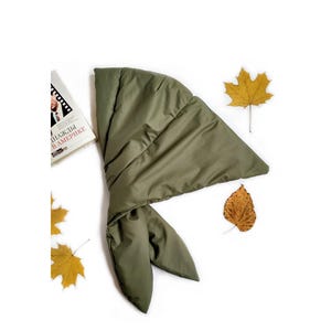 May include: An olive green quilted scarf with a smooth, padded texture and a tapered, pointed end. The scarf is positioned next to a book with the Russian text "Однажды в Америке" visible on the cover, accompanied by several scattered yellow and brown autumn leaves.