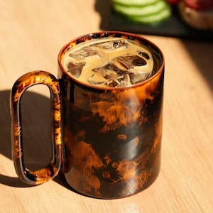 May include: A ceramic coffee mug with a unique marbled design in shades of brown and black, filled with iced coffee. The mug has a large, curved handle. The background includes sliced cucumbers and tomatoes.